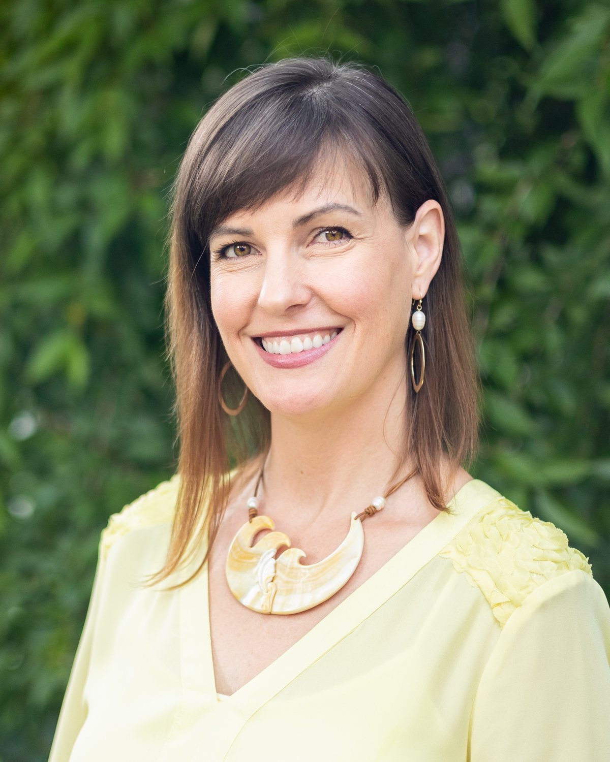 Ashley Oros in a yellow blouse, smiling against green foliage.