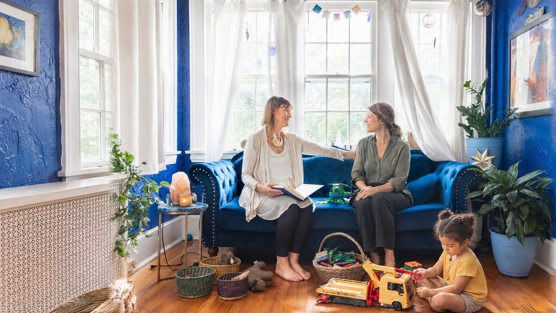Ashley Oros beside a parent on a blue velvet sofa, with a child playing nearby.